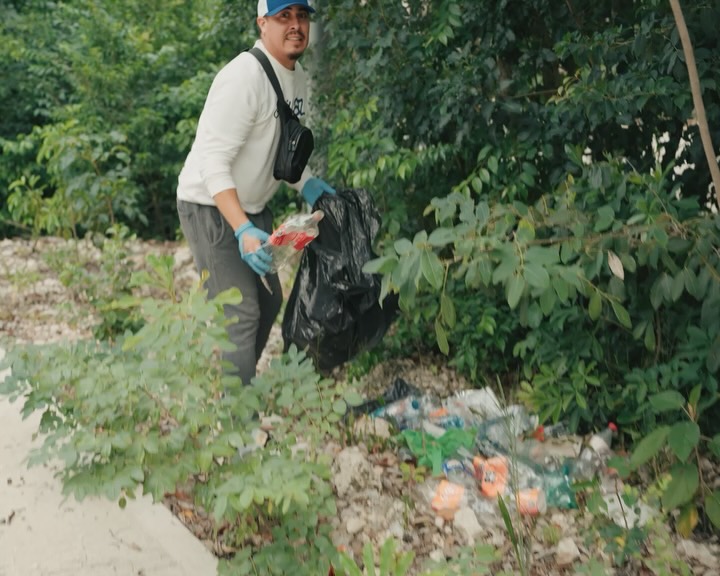 A volunteer in gloves collects litter into a black bag along a jungle roadside.