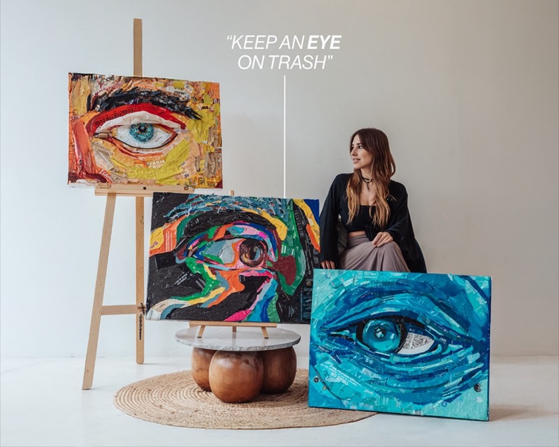 Artist Ana Pau sits beside three colourful eye artworks made from waste materials.